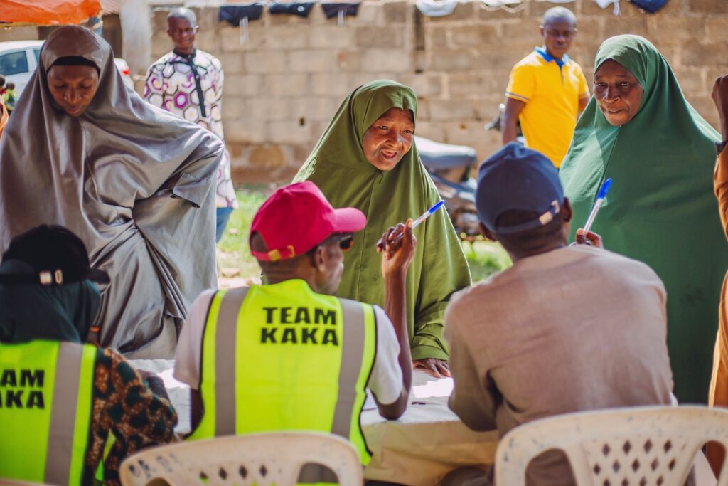 Men and women seated outdoors in a community consultation meeting in Kaduna, some holding pens and papers
