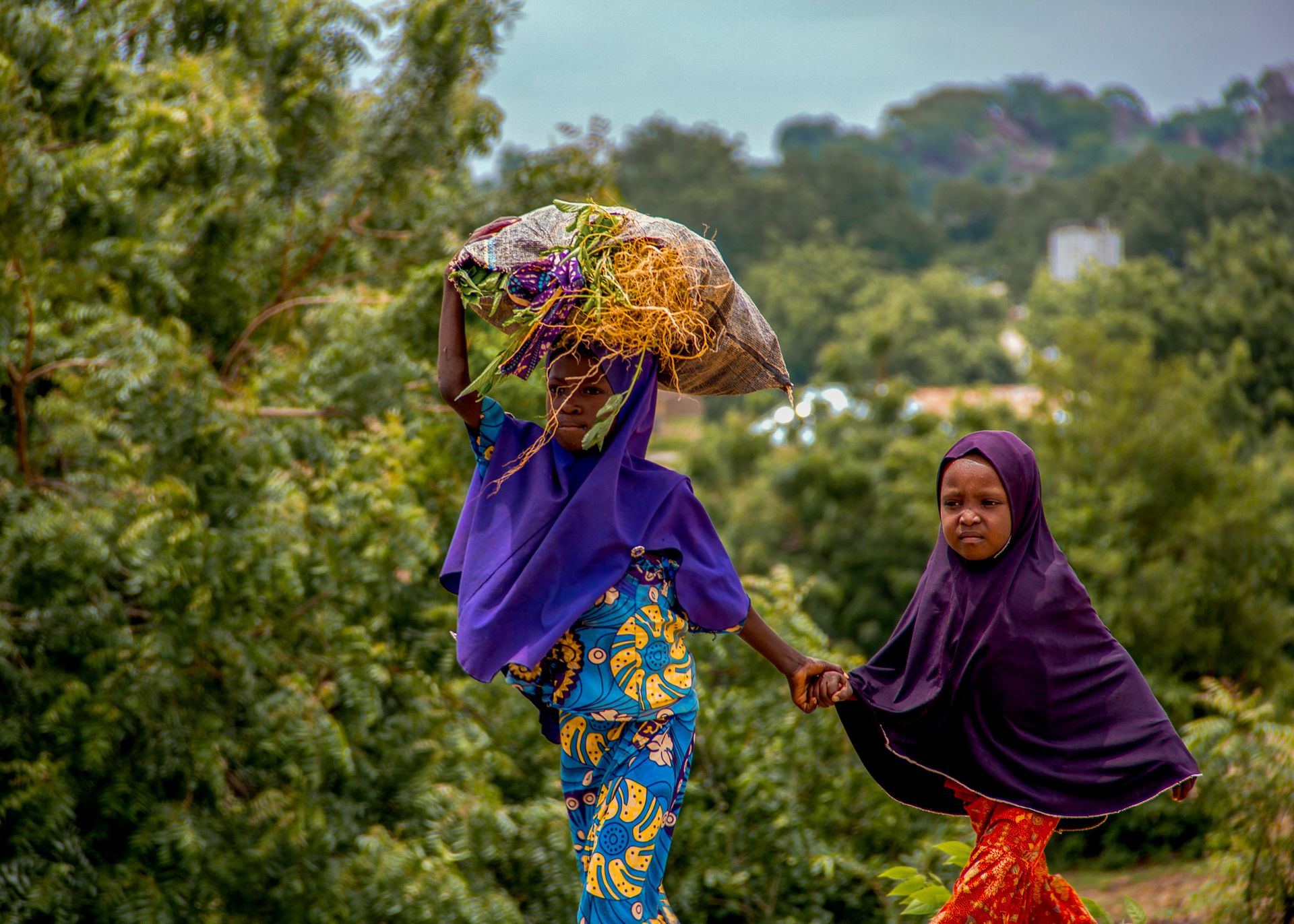 A mother in bright traditional dress walks a rural path with her child, carrying harvest