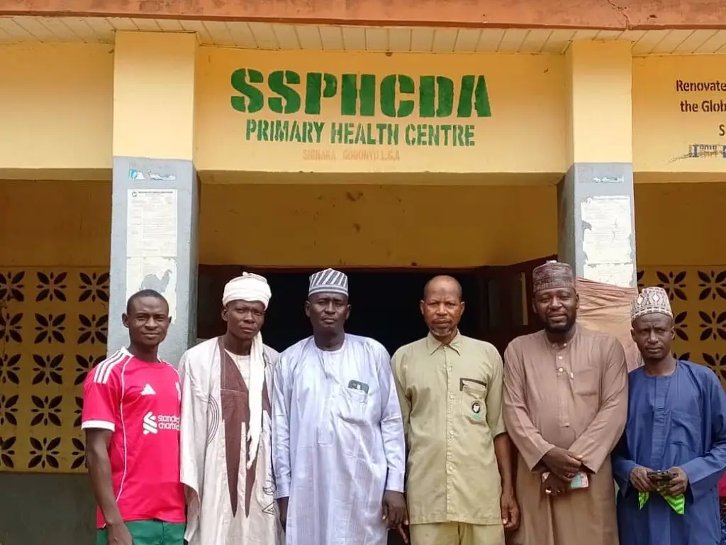 LHAAT members standing under the SSPHCDA Primary Health Centre sign in Goronyo, Sokoto